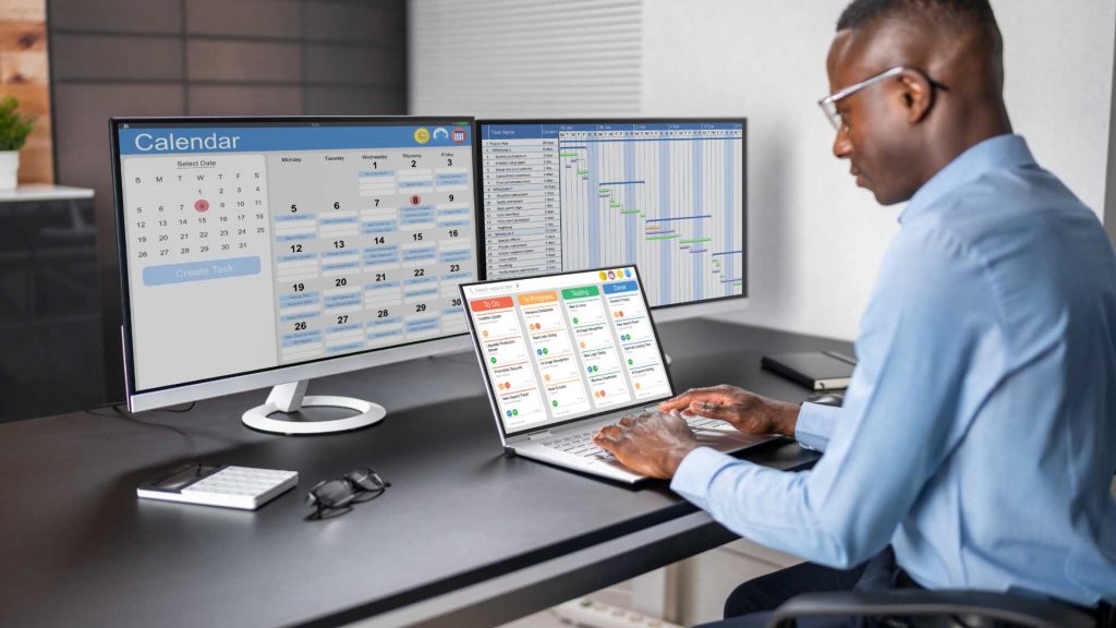 Homem de negócios trabalhando em múltiplos dispositivos eletrônicos com agenda, calendário, planilha e smartphone em escritório moderno fazendo gestão de projetos
